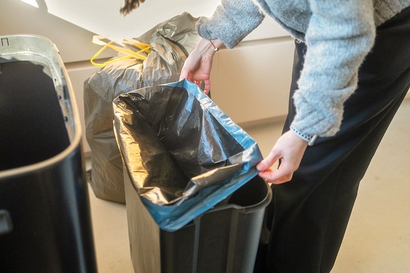 Anonymous person putting a new trash bag in a black garbage bin. You can see a full bag of trash in the background.