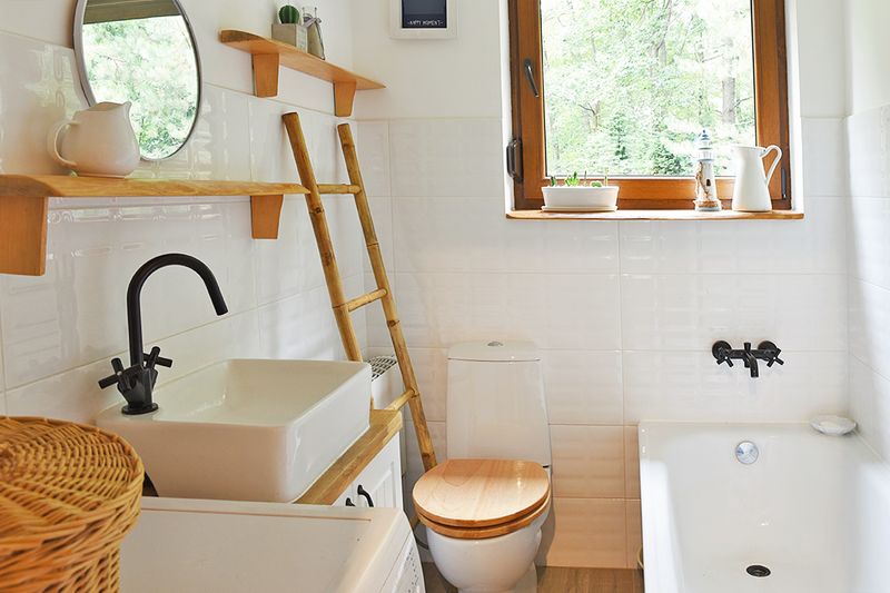 A mostly-white bathroom with wood accents, such as open shelves, a bamboo towel ladder, and a toilet seat lid.