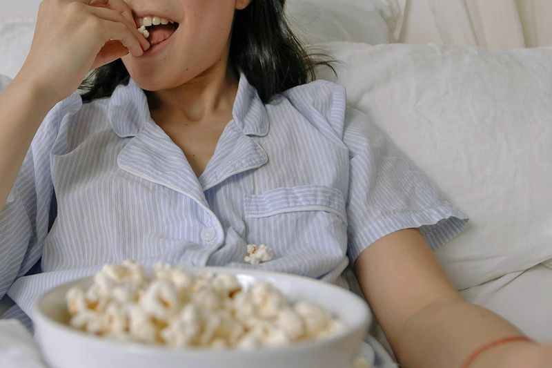Anonymous woman in pajamas eating a bowl of popcorn in bed.