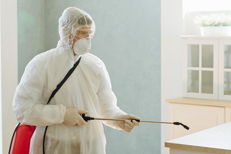 Man in a white hazmat suit spraying a chemical from a red tank in a kitchen.
