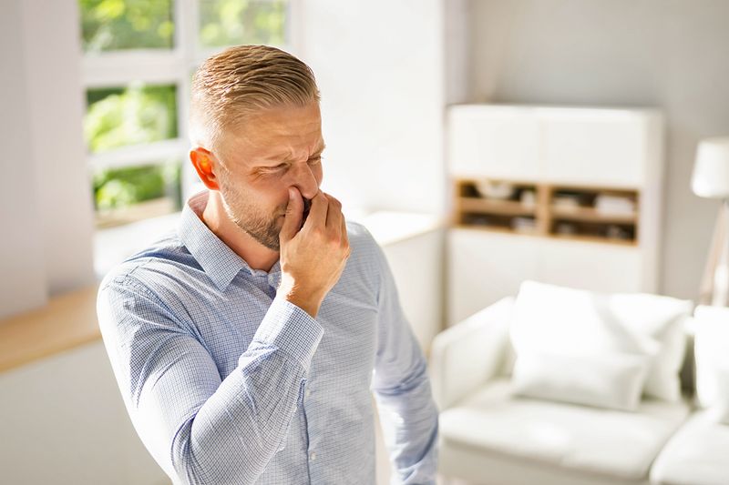 A blond man in a button-down shirt plugging his nose and crinkling his eyes at a smell in the house he's standing in.