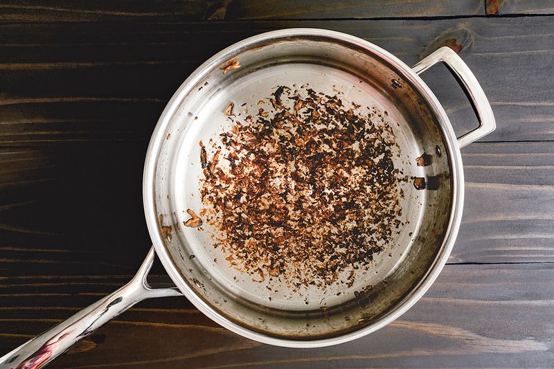 A stainless steel skillet with burnt-on residue at the bottom. The skillet is placed on a wood countertop.