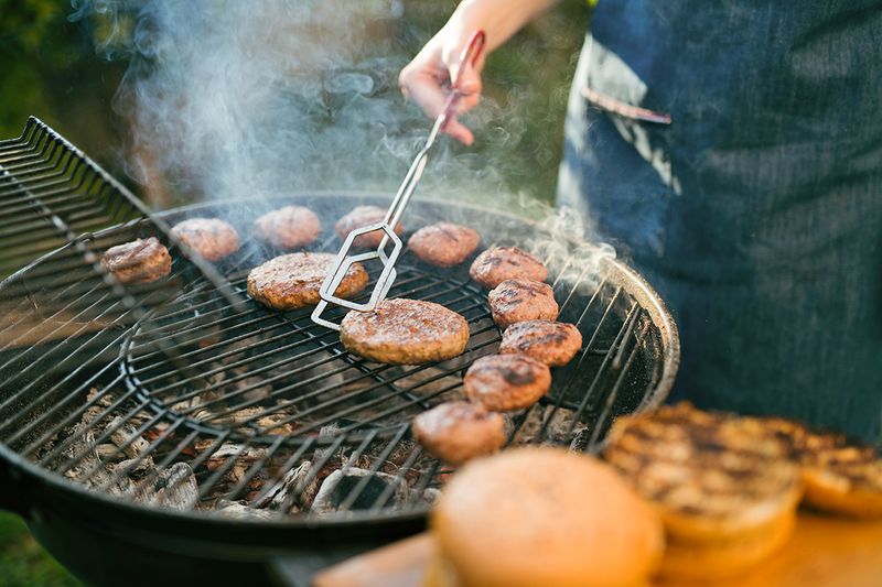 Anonymous person flipping burgers on a round, charcoal grill outdoors.