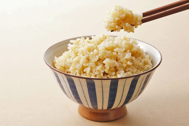 A small, blue and white striped ceramic bowl full of brown rice. A pair of chopsticks is holding up a bite of it.