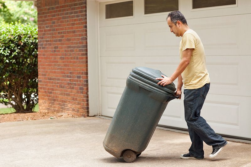 A middle-aged man wheeling an outdoor trash can to the curb, past a home's garage.