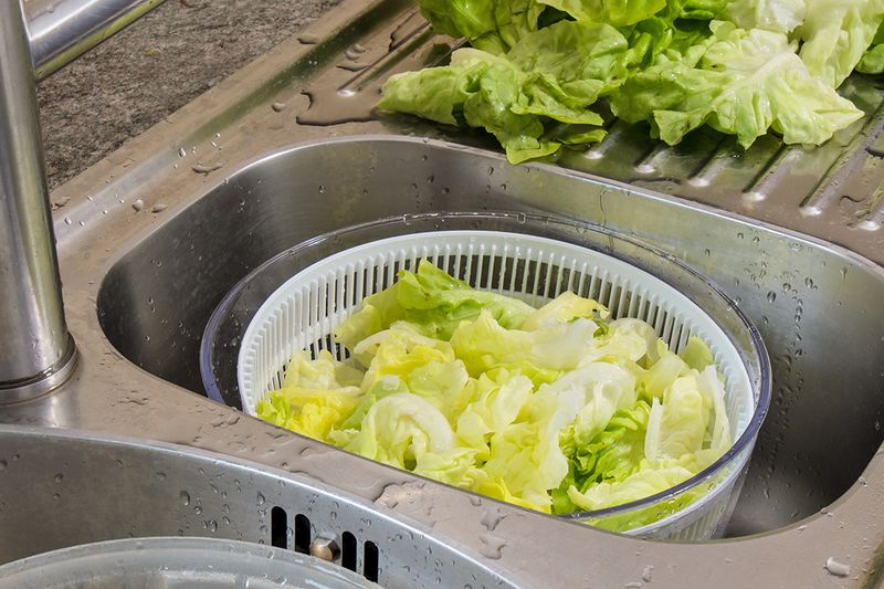 A colander full of chopped lettuce in one half of a divided stainless steel sink.