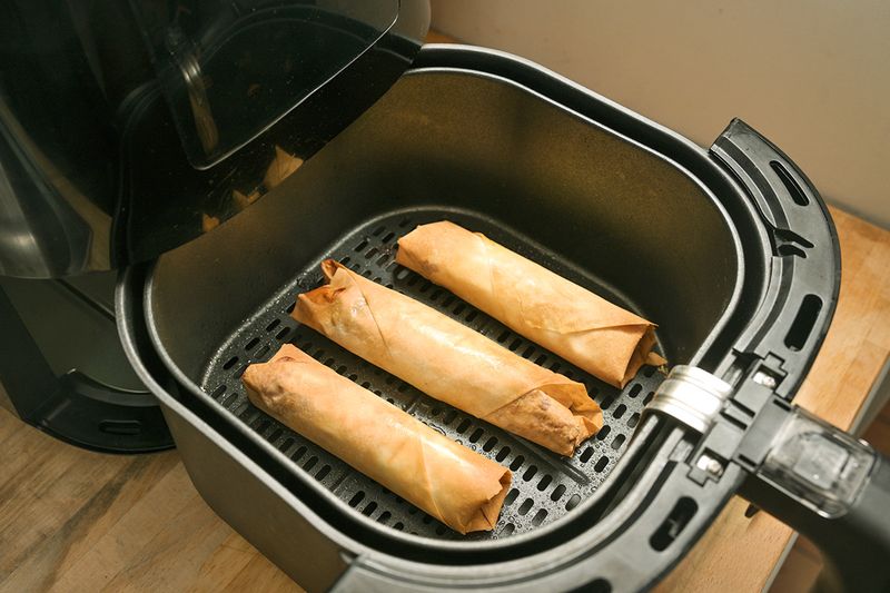 An open air fryer showing the basket with three crisped-up spring rolls.