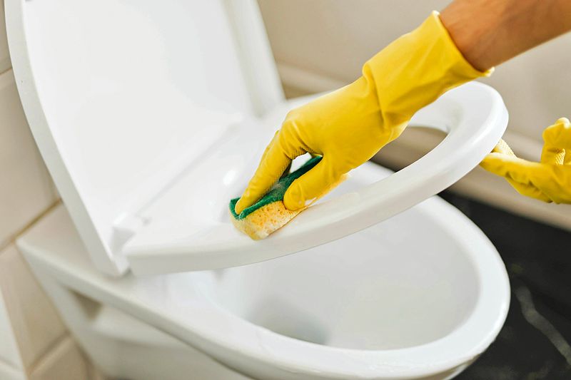 Anonymous person wearing yellow gloves cleaning a raised toilet seat with a sponge.