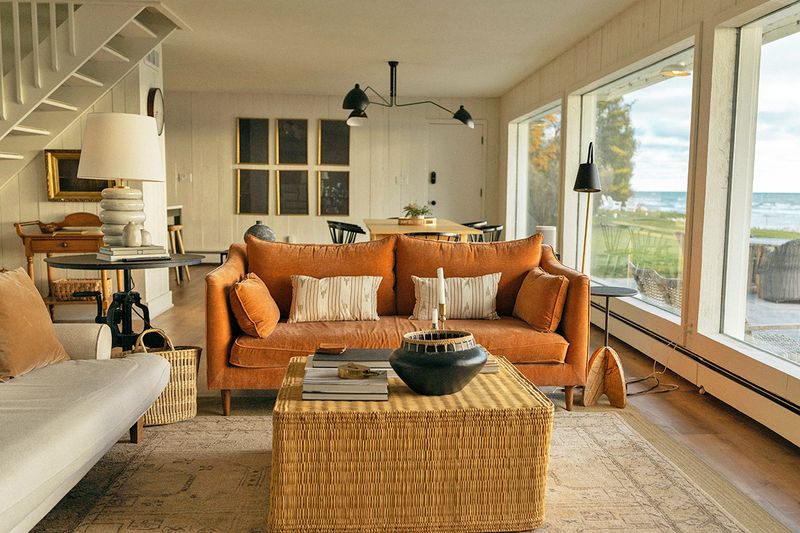 A lake house living room with an orange sofa, wicker coffee table, and paneled white walls.