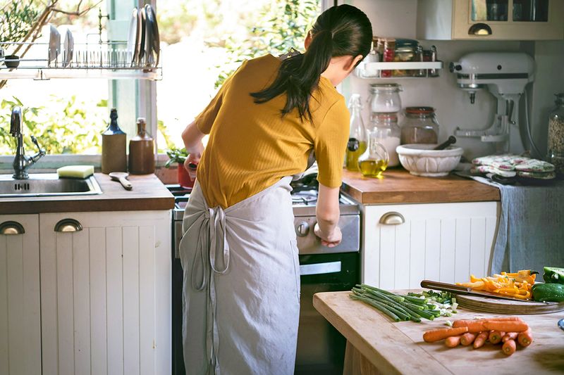 A woman lighting a gas stove. Behind her on a kitchen island are several vegetables ready for chopping on a cutting board.