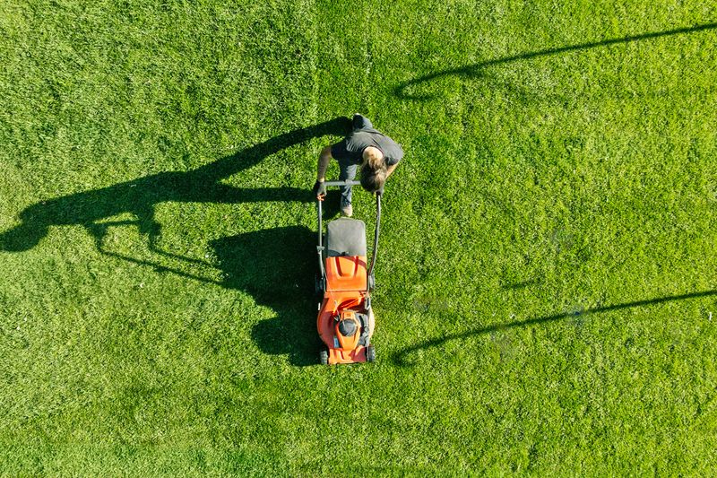 An overhead shot of an anonymous person pushing an orange lawn mower over green grass.