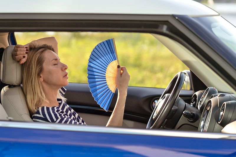 A woman in a blue car with all the windows rolled down, fanning herself with a folding fan.