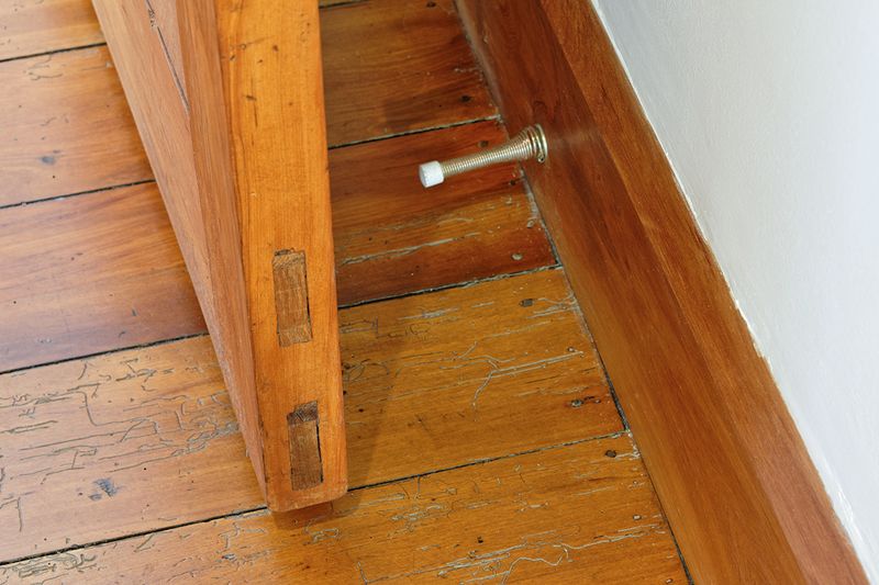 A metal, spring-loaded doorstop attached to a wooden baseboard.