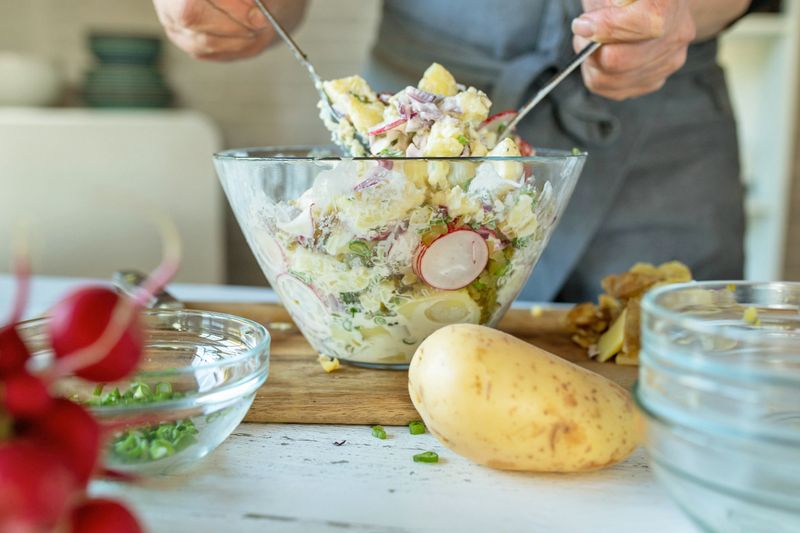An anonymous person tossing a potato salad with mayonnaise, radishes, green onion, red onion, and other ingredients.