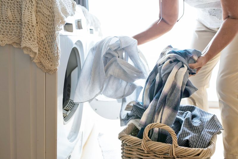 Anonymous person loading dirty clothes into a washing machine from a wicker basket.