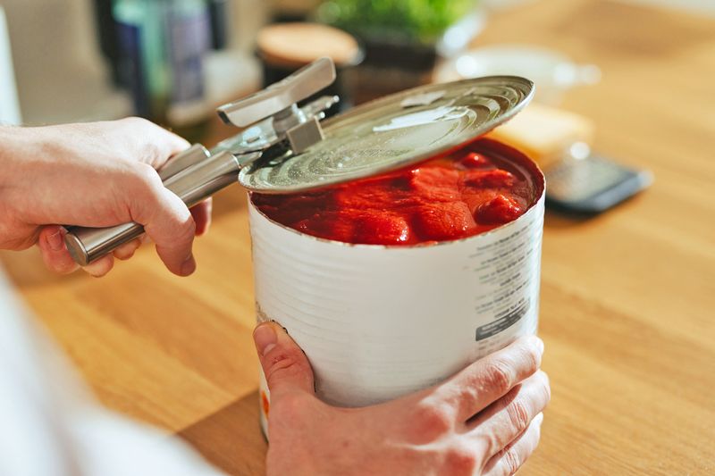 Anonymous person using a can opener to open a large can of tomatoes.