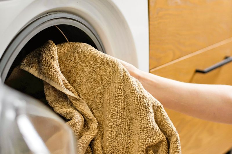 Anonymous person placing a tan bath towel in a front-loading washing machine.
