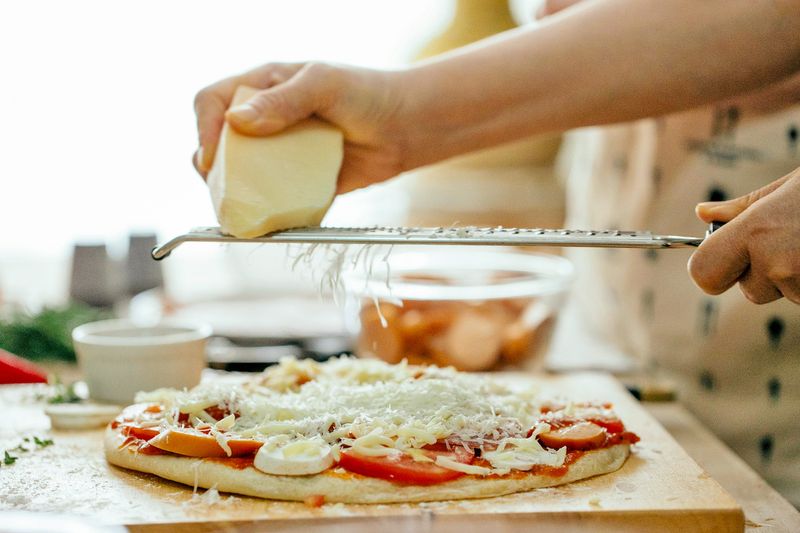 Anonymous person grating a block of mozzarella onto a pizza before baking.