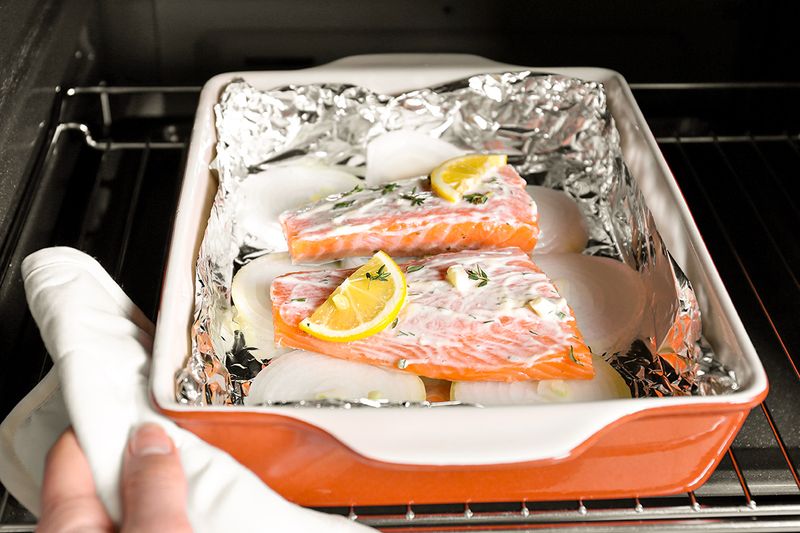 Anonymous person placing a baking dish with two pieces of salmon covered in mayonnaise, lemon slices, and fresh herbs in the oven.
