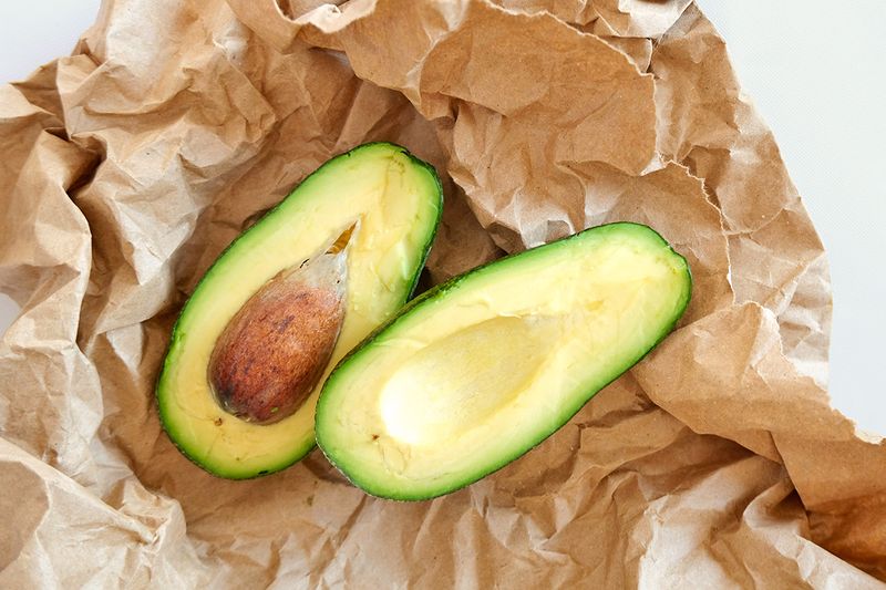 Two cut halves of a ripe avocado sitting on a crumpled paper bag.