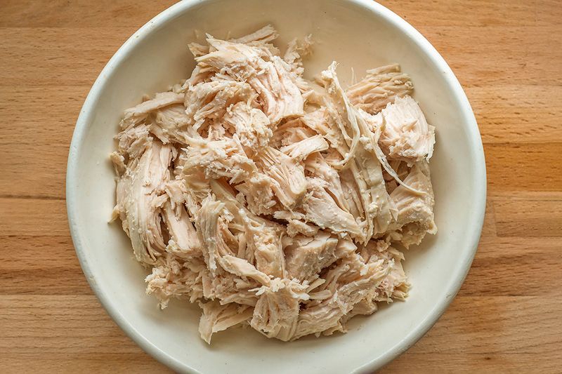 A white ceramic bowl full of shredded rotisserie chicken on a wooden surface.