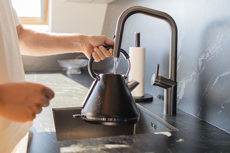 Anonymous person filling up a black matte electric kettle at a kitchen faucet.