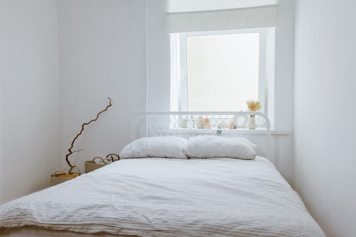 White bed in small white room with window behind bed frame.