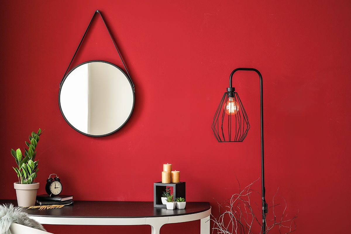 Room with red wall and black wire lamp; round, hanging black mirror; and black table with plants, candles, and an alarm clock.