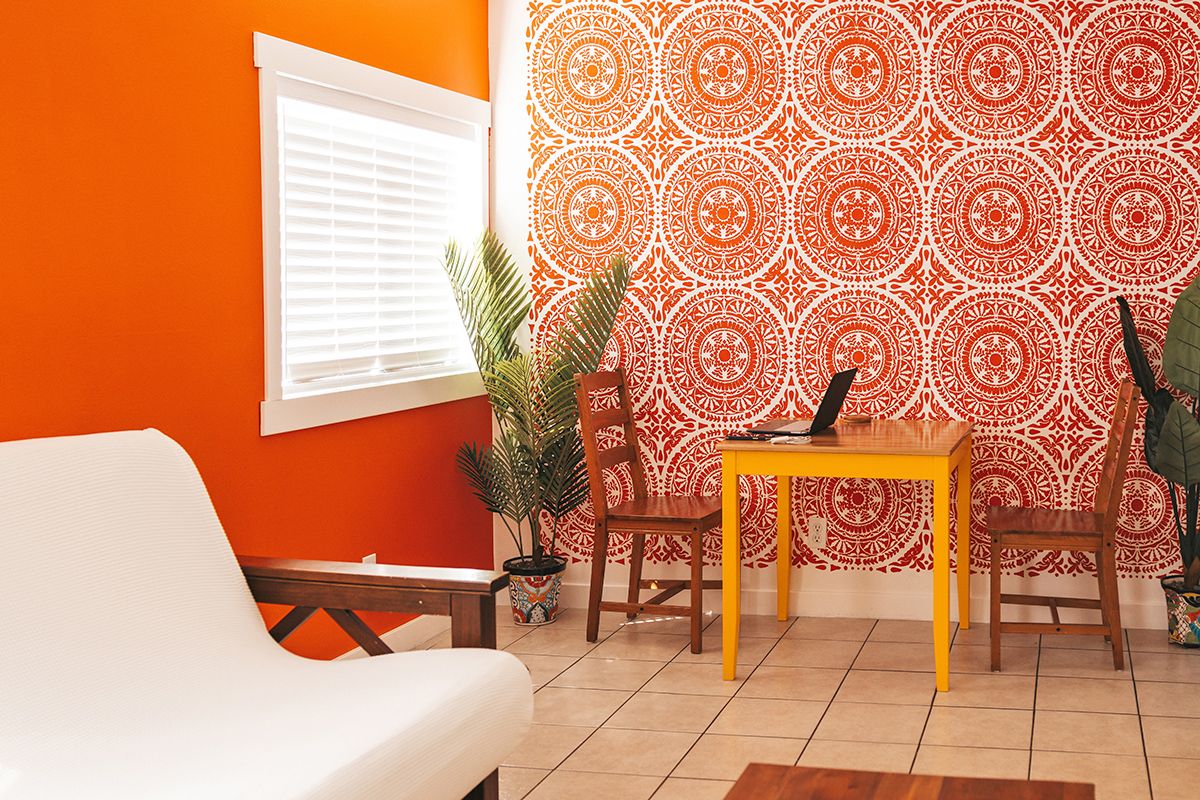Room with one plain orange wall with a window and one patterned white and orange wall, chairs, plants, and a table with a laptop.