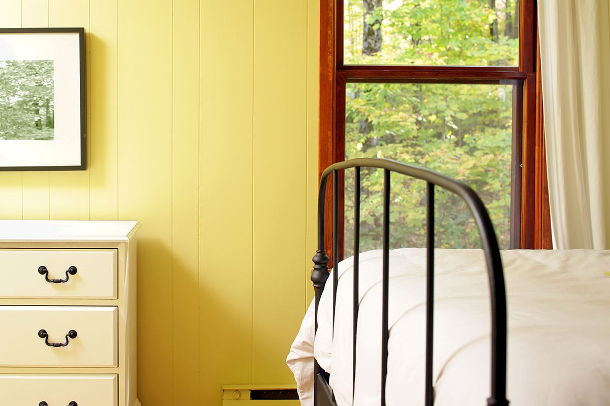 Part of bedroom with yellow wall, window, end of bed in black iron bed frame, dresser, and hanging picture in black frame.