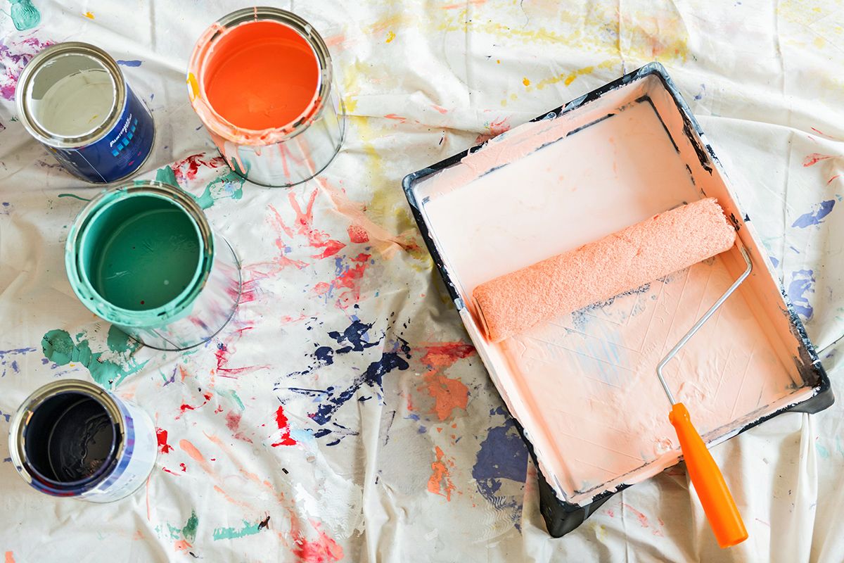 Roller in tray of light orange paint next to cans of white, black, green, and orange paint on paint-splattered sheet.