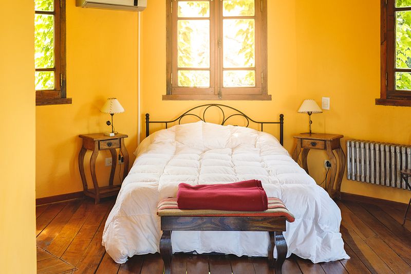Bed with white duvet in small room with warm yellow walls with three windows with sunlight streaming in and a table at the foot of the bed with a folded red blanket on it.